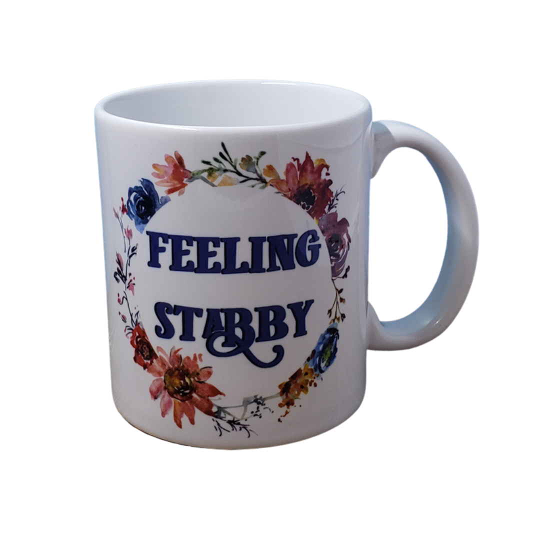White coffee mug decorated with a floral wreath. Inside the wreath the words "feeling stabby" are written in all caps.